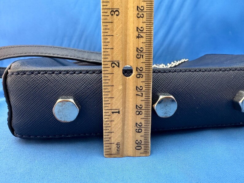 Puede incluir: Un bolso de mano azul marino con herrajes plateados. El bolso mide aproximadamente 5 cm de altura. Se coloca una regla de madera junto al bolso para medir.
