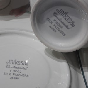 May include: Close-up of the bottom of a white Mikasa Continental F 3003 Silk Flowers teacup and saucer. The text "Mikasa Continental F 3003 Silk Flowers Japan" is printed in gray on the bottom of the cup and saucer.