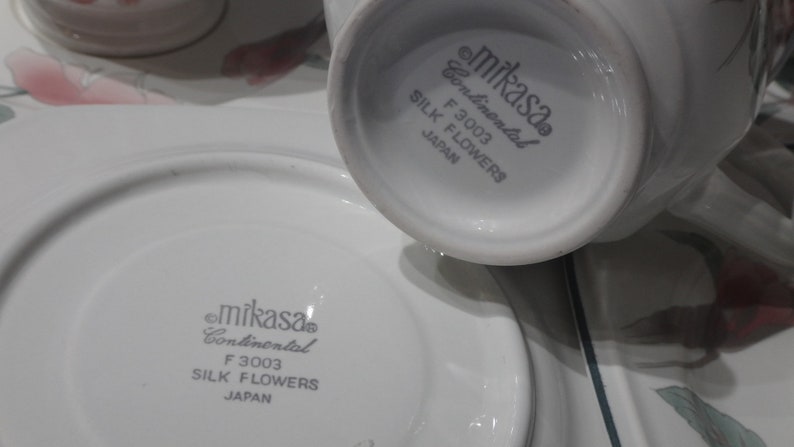 May include: Close-up of the bottom of a white Mikasa Continental F 3003 Silk Flowers teacup and saucer. The text "Mikasa Continental F 3003 Silk Flowers Japan" is printed in gray on the bottom of the cup and saucer.