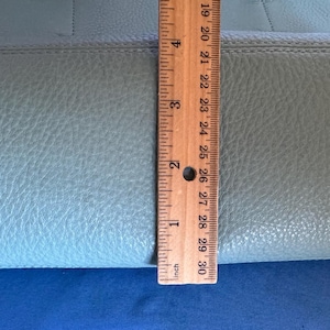 May include: A light blue leather handbag with a textured surface. A wooden ruler is placed vertically against the bag, showing a measurement of approximately 10 inches.