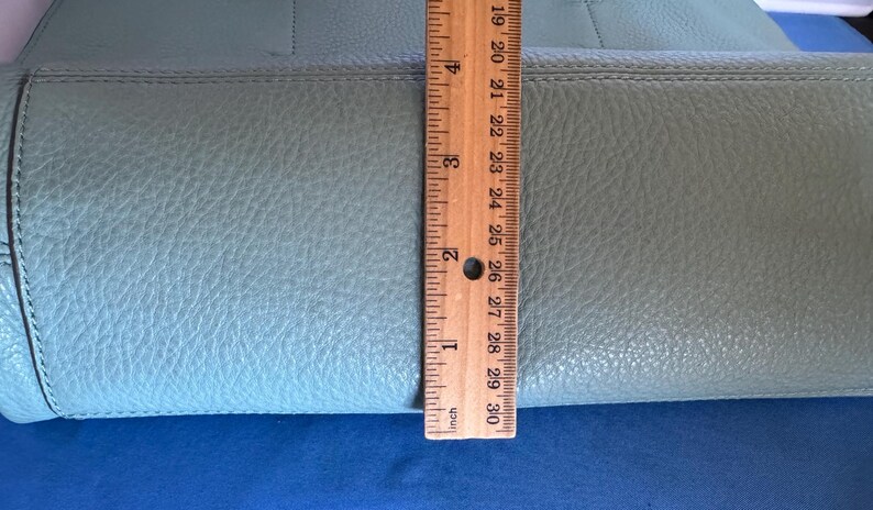 May include: A light blue leather handbag with a textured surface. A wooden ruler is placed vertically against the bag, showing a measurement of approximately 10 inches.