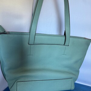 May include: A light teal leather tote bag with two long handles. The bag has a rectangular shape with a front pocket and a textured surface. The handles are stitched to the top of the bag.