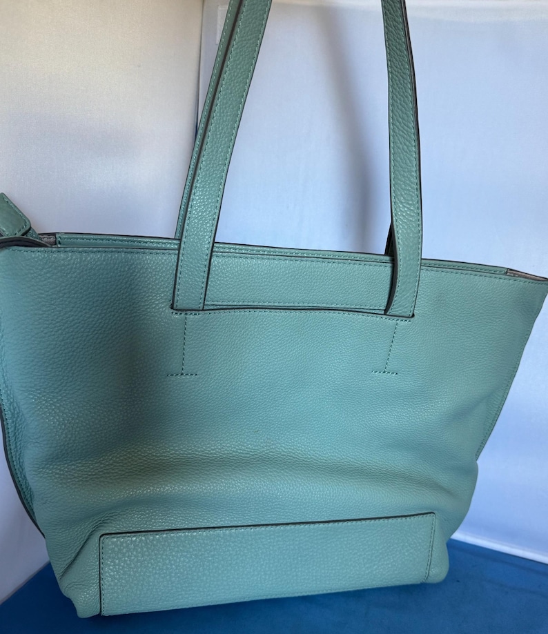 May include: A light teal leather tote bag with two long handles. The bag has a rectangular shape with a front pocket and a textured surface. The handles are stitched to the top of the bag.