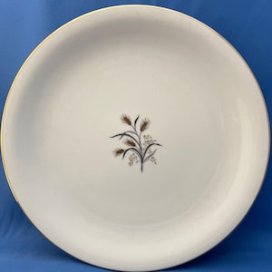 May include: A white ceramic plate with a gold rim and a wheat stalk design in the center. The wheat is brown and black, with small white flowers. The plate is round and has a simple, elegant design.