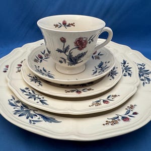 May include: A cream-colored ceramic tea set consisting of a teacup, a saucer, a small plate, and a large dinner plate. Each piece features a delicate floral pattern with dark blue leaves, red flowers, and small yellow berries on the edges.