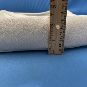 May include: A white leather handbag with a textured surface. The bag is approximately 2 inches (5 cm) wide, as measured by a wooden ruler. The bag has a curved shape and a shoulder strap.