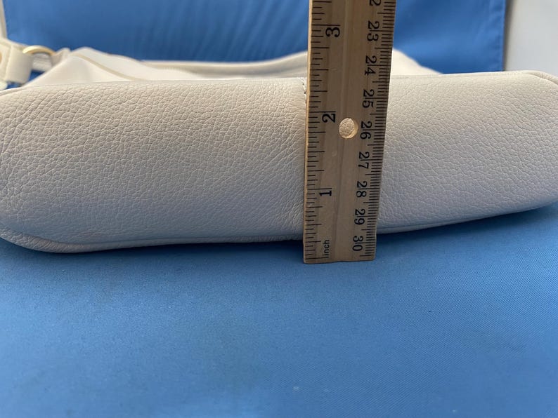 May include: A white leather handbag with a textured surface. The bag is approximately 2 inches (5 cm) wide, as measured by a wooden ruler. The bag has a curved shape and a shoulder strap.