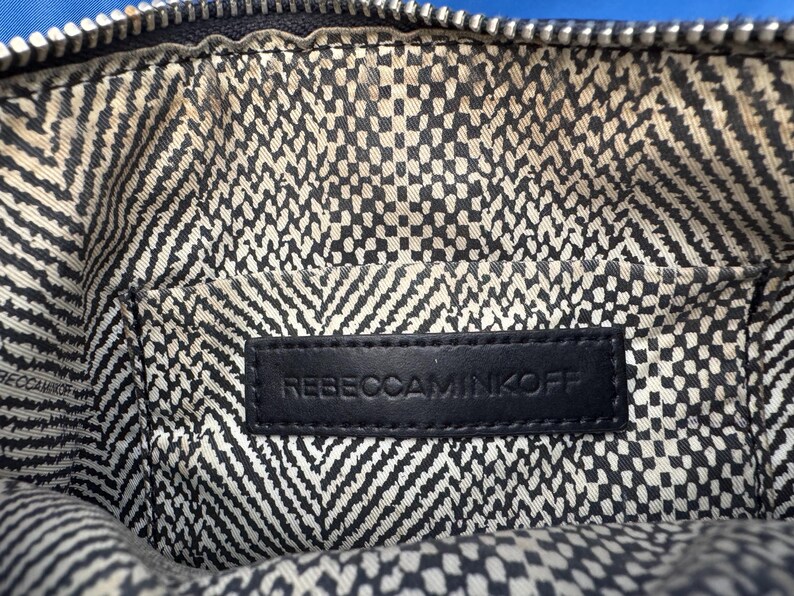 Puede incluir: Primer plano del interior de un bolso negro. El forro presenta un patr&oacute;n geom&eacute;trico en blanco y negro. Se ve una etiqueta de cuero negro con el texto en relieve "REBECCAMINKOFF".