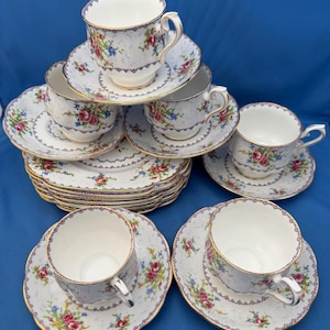 May include: A collection of vintage teacups and saucers. The white porcelain cups and saucers feature a floral pattern with red, pink, and blue flowers. The rims are trimmed with gold and a blue geometric design. The set includes multiple cups and saucers.