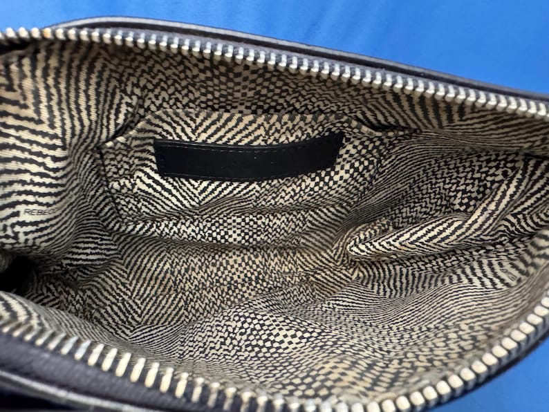 Puede incluir: Vista interior de una bolsa negra con cremallera y forro estampado en blanco y negro. El forro presenta una mezcla de patrones de chevron y cuadrados peque&ntilde;os. Una etiqueta negra est&aacute; cosida en el interior de la bolsa.