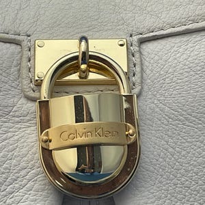 May include: Close-up of a white leather handbag with a gold-colored lock. The lock features the brand name "Calvin Klein" in gold lettering. The bag's leather has a textured appearance.