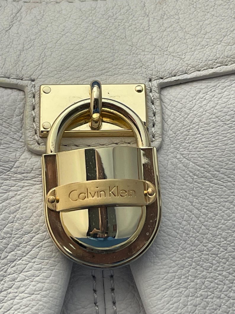 May include: Close-up of a white leather handbag with a gold-colored lock. The lock features the brand name "Calvin Klein" in gold lettering. The bag's leather has a textured appearance.