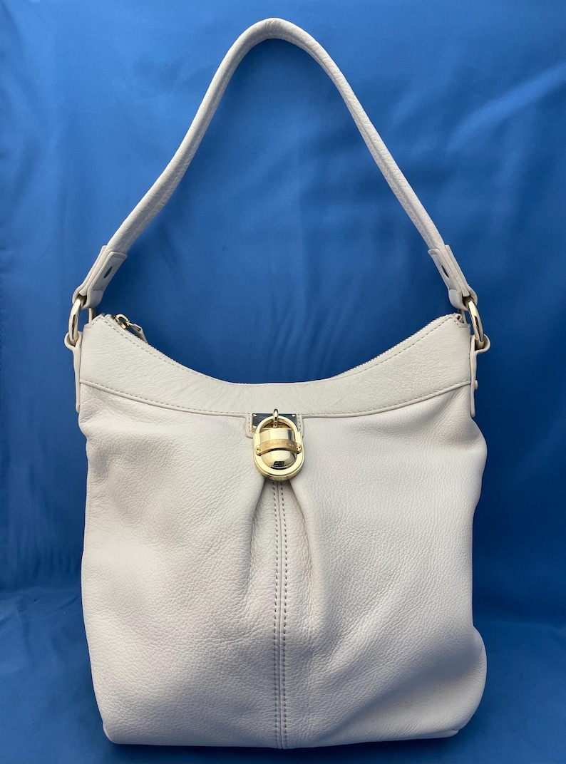 May include: A light gray leather shoulder bag with a single strap. The bag has a gold-colored clasp and zipper. The bag is made of soft leather and has a simple, elegant design.
