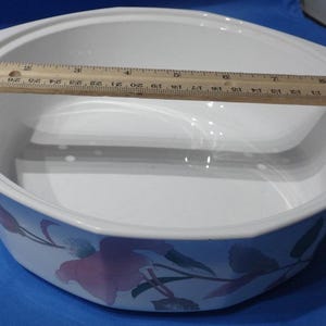 May include: White ceramic baking dish with a floral design featuring pink and green flowers. The dish has a removable lid and a built-in steamer insert.