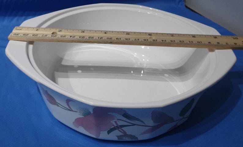 May include: White ceramic baking dish with a floral design featuring pink and green flowers. The dish has a removable lid and a built-in steamer insert.