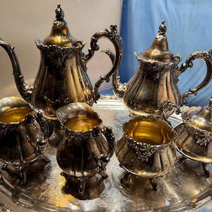 May include: A silver-plated tea service set consisting of a large teapot, a smaller teapot, a creamer, a sugar bowl, and a waste bowl, all featuring ornate floral embossed details and curved handles, arranged on a large oval silver serving tray.