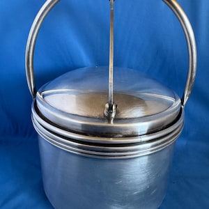 Könnte beinhalten: Ein silberfarbener Metalleiskübel mit rundem Körper und Deckel. Der Eimer hat einen großen, gebogenen Griff und einen kleineren Griff am Deckel. Das Design ist schlicht und elegant, geeignet zum Servieren von gekühlten Getränken.