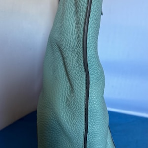 May include: A light teal handbag with a textured leather exterior. The bag has a rounded shape and a dark trim along the edges. The strap is a matching teal color.