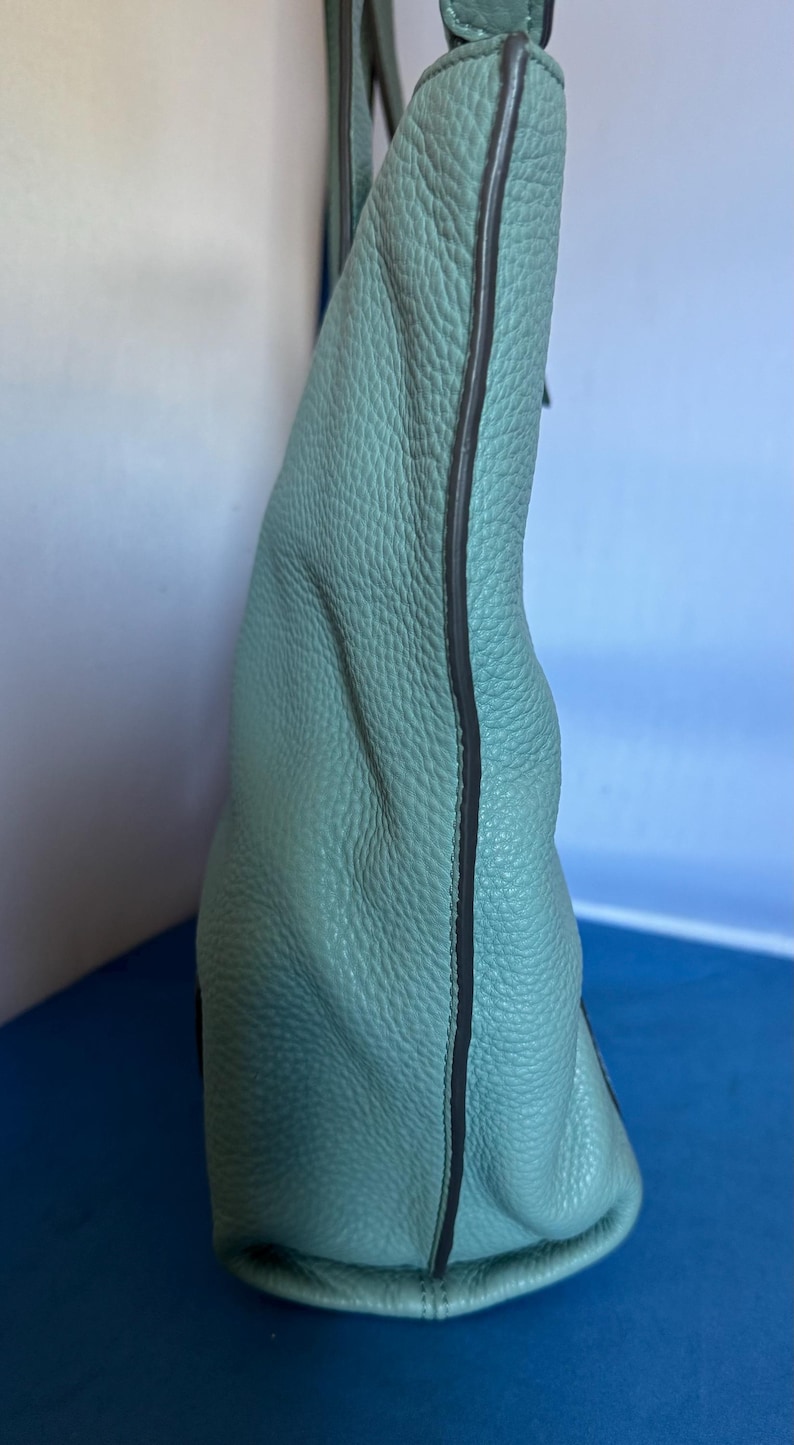 May include: A light teal handbag with a textured leather exterior. The bag has a rounded shape and a dark trim along the edges. The strap is a matching teal color.