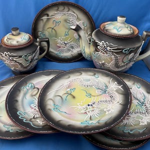 Op de afbeelding: Een zwart-wit theeservies met drakenmotief met een theepot, suikerpot en borden. De borden en het theeservies hebben een drakenontwerp met kleurrijke accenten. De set bevat een theepot, een suikerpot en zes borden.