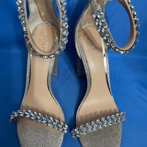 May include: A pair of silver glitter heeled sandals with ankle straps adorned with crystal embellishments. The sandals feature the brand name "J.Crew" on the insole.