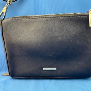 Puede incluir: Un bolso bandolera azul marino con cremallera plateada y correa de cadena. El bolso tiene forma rectangular con una superficie texturizada y un peque&ntilde;o logotipo plateado. El fondo es azul liso.