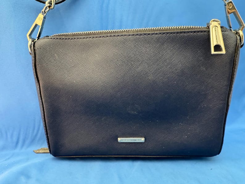 Puede incluir: Un bolso bandolera azul marino con cremallera plateada y correa de cadena. El bolso tiene forma rectangular con una superficie texturizada y un peque&ntilde;o logotipo plateado. El fondo es azul liso.