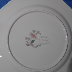 May include: White ceramic dinner plate with a scalloped edge. The plate features a floral design with pink flowers and green leaves. The back of the plate has the text "Mikasa Continental Silk Flowers" and "Oven to Table Dishwasher Safe for Microwave Oven Japan."