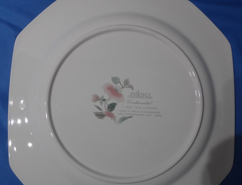 May include: White ceramic dinner plate with a scalloped edge. The plate features a floral design with pink flowers and green leaves. The back of the plate has the text "Mikasa Continental Silk Flowers" and "Oven to Table Dishwasher Safe for Microwave Oven Japan."