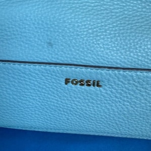May include: Close-up of a light blue leather bag with the brand name "FOSSIL" in gold lettering. The bag has a textured surface and a dark blue accent line.