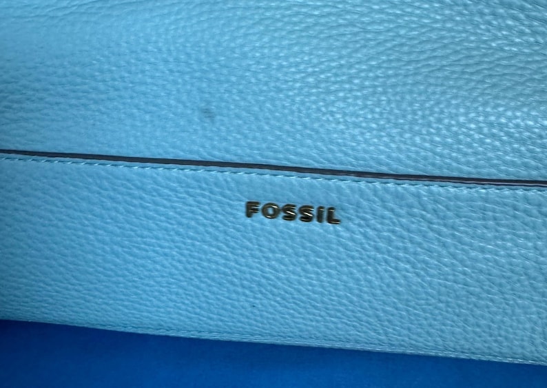 May include: Close-up of a light blue leather bag with the brand name "FOSSIL" in gold lettering. The bag has a textured surface and a dark blue accent line.