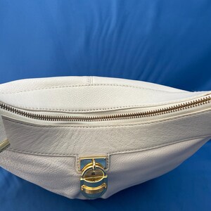 May include: A white leather shoulder bag with a curved shape. The bag features a gold-toned zipper and a decorative clasp. The strap is also white leather. The bag is set against a blue background.