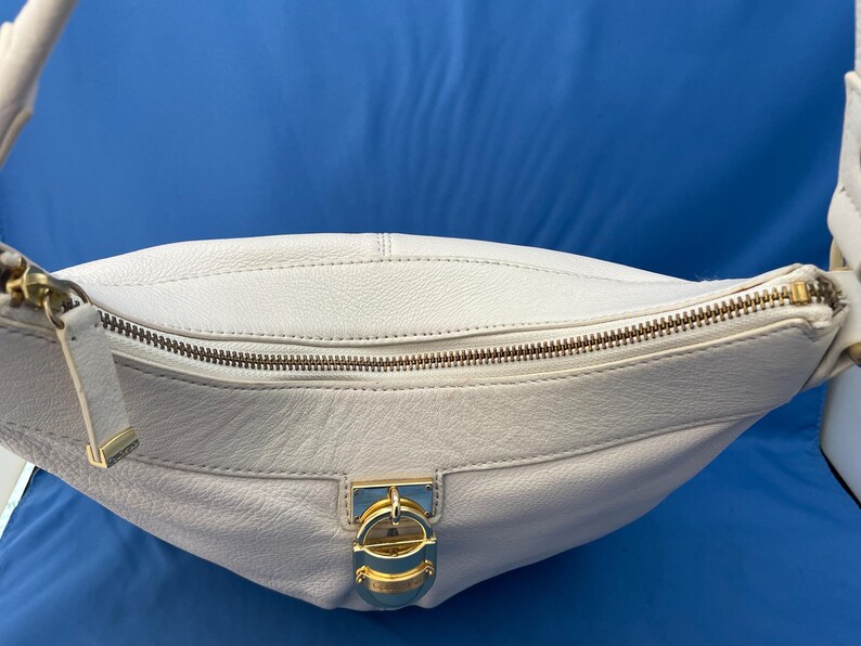 May include: A white leather shoulder bag with a curved shape. The bag features a gold-toned zipper and a decorative clasp. The strap is also white leather. The bag is set against a blue background.