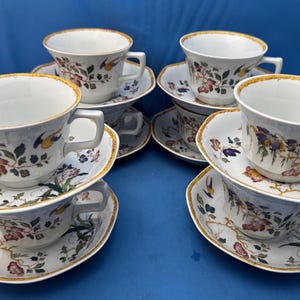 May include: Six white teacups and saucers with floral and bird designs. The cups and saucers have a gold trim. The floral pattern includes red, purple, and green accents. The teacups are stacked on top of the saucers.