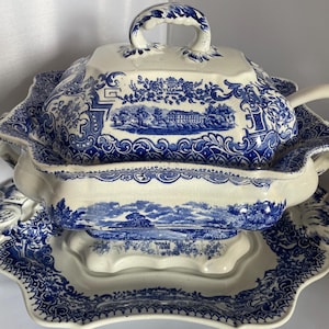 Op de afbeelding: Een blauw-witte keramische soepterrineset. De set bevat een terrine met deksel en handvat, een pollepel en een decoratieve basis. De terrine en de basis hebben een blauw landschapsontwerp. De set is waarschijnlijk vintage.