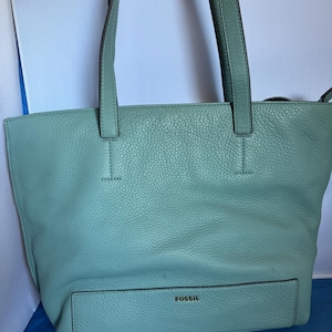 May include: A light teal leather tote bag with two handles and a front pocket. The bag has a pebbled texture and the brand name "FOSSIL" is embossed on the front pocket.