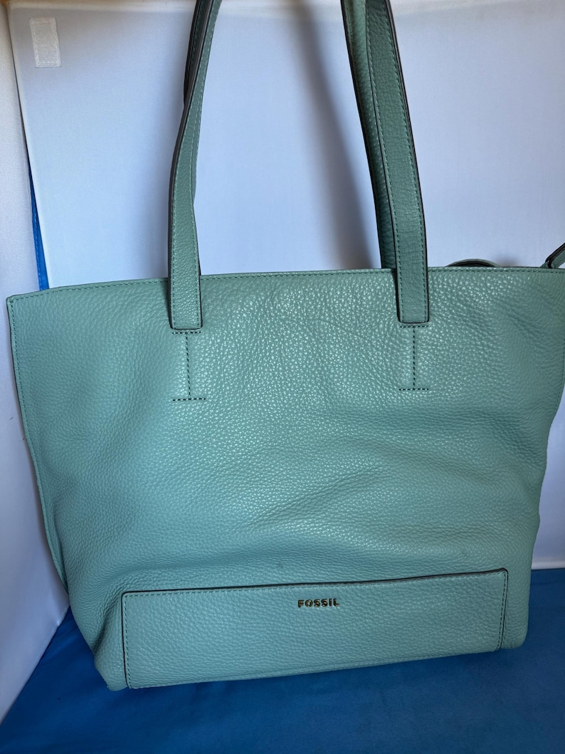 May include: A light teal leather tote bag with two handles and a front pocket. The bag has a pebbled texture and the brand name "FOSSIL" is embossed on the front pocket.