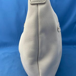 May include: A light gray leather shoulder bag with a rounded shape. The bag has a textured surface and a strap with a gold-colored button detail. The background is a solid blue.