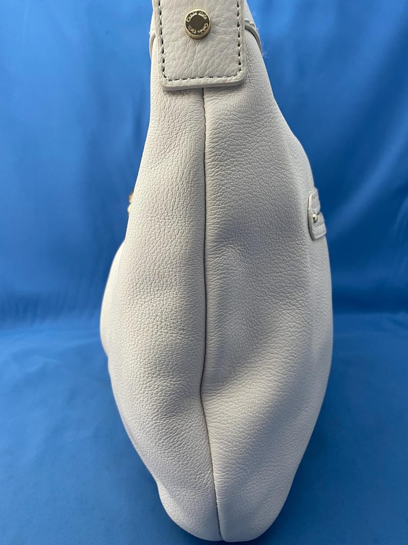 May include: A light gray leather shoulder bag with a rounded shape. The bag has a textured surface and a strap with a gold-colored button detail. The background is a solid blue.