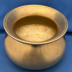 Könnte beinhalten: Ein antiker Messingtopf mit breitem, ausgestelltem Rand und rundem Körper. Der Topf weist Gebrauchsspuren auf, mit Flecken und Unvollkommenheiten auf der Oberfläche. Der offene Innenraum deutet darauf hin, dass er zum Kochen oder Aufbewahren verwendet wurde.