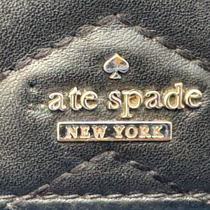 Może przedstawiać: Zbliżenie czarnej, pikowanej powierzchni ze sk&oacute;ry z logo Kate Spade New York. Logo zawiera nazwę marki w złotych literach, symbol pik i słowa "NEW YORK" w prostokątnej ramce.