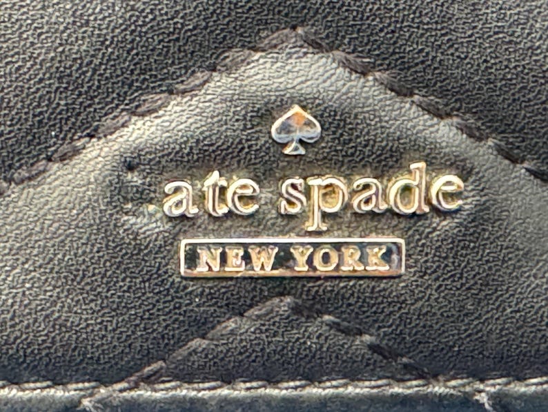 Może przedstawiać: Zbliżenie czarnej, pikowanej powierzchni ze sk&oacute;ry z logo Kate Spade New York. Logo zawiera nazwę marki w złotych literach, symbol pik i słowa "NEW YORK" w prostokątnej ramce.