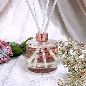 Könnte beinhalten: Eine klare Glasdiffuserflasche mit einem roségoldenen Deckel und weißen Diffusorsticks. Die Flasche ist mit einer klaren Flüssigkeit und rosa Blüten gefüllt. Der Diffusor steht auf einer weißen Oberfläche.