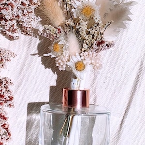 May include: A clear glass diffuser bottle with a rose gold lid and white diffuser sticks. The bottle is decorated with a bouquet of dried flowers, including white daisies, beige pampas grass, and pink dried flowers.