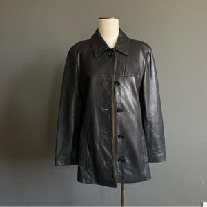 May include: A black leather jacket with a collared neckline and button-down closure. The jacket is displayed on a mannequin and has a classic, tailored design. The jacket has a mid-length cut and is suitable for various occasions.