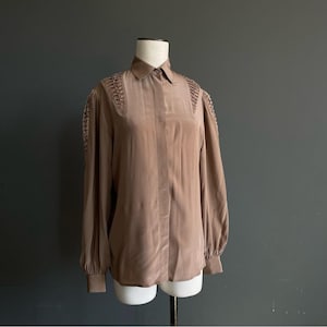 May include: A long-sleeved, button-down blouse in a muted brown hue. The blouse features a classic collar and decorative detailing on the shoulders. The sleeves are gathered at the cuffs, and the fabric has a subtle sheen.
