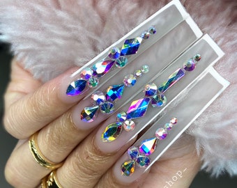 May include: Close-up of long, clear acrylic nails with iridescent rhinestones arranged in a decorative pattern. The nails are on a hand wearing gold rings. The image includes the text "@bbnailshop_"