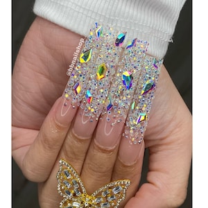 May include: Long, clear acrylic nails with a butterfly ring. The nails are decorated with iridescent rhinestones and small white beads.