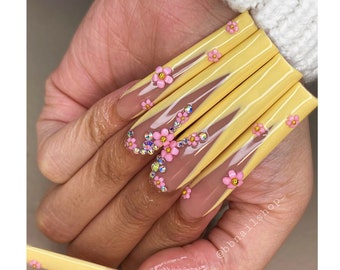 May include: Close-up of a hand with long, yellow and nude nails decorated with pink flowers and rhinestones. The nails are shaped into a long, pointed shape. The hand is holding a yellow nail file. The Instagram handle @bbnailshop is visible in the bottom right corner of the image.