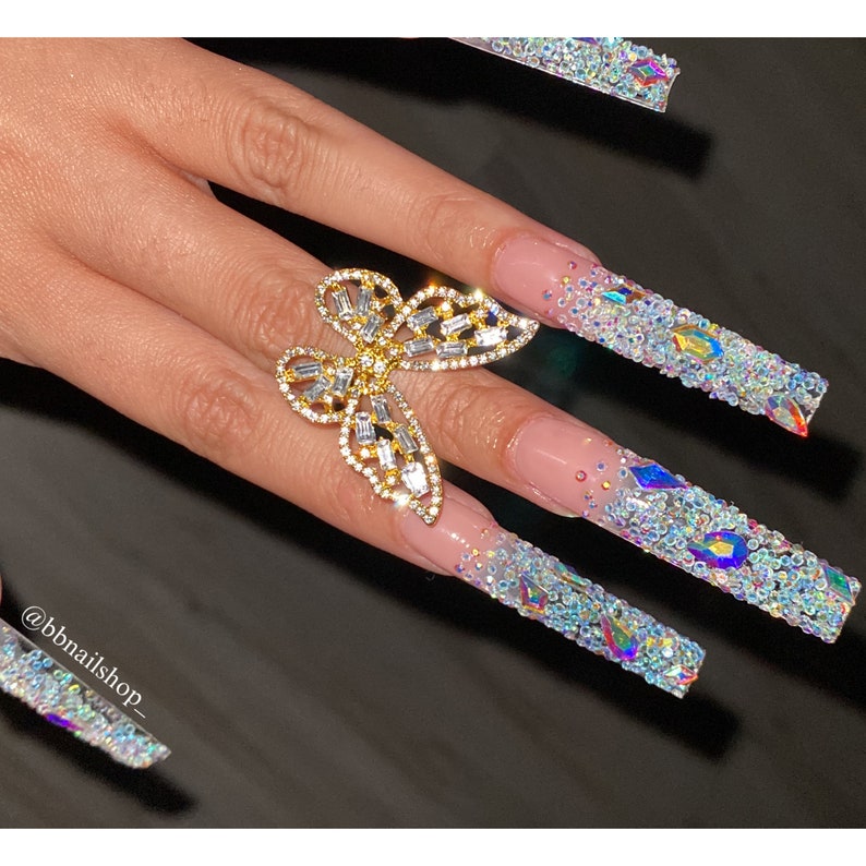 May include: Long, clear, acrylic nails with a butterfly ring. The nails are decorated with iridescent rhinestones and glitter.